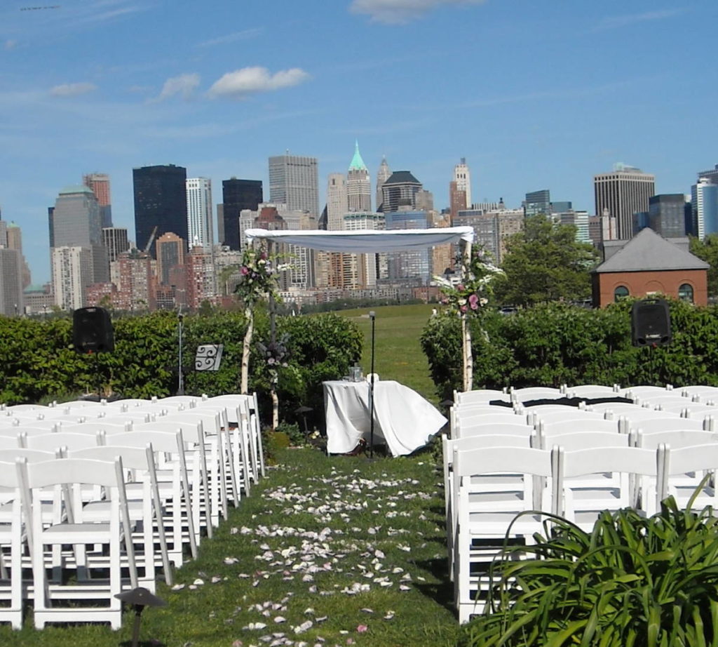 Wind tugs at the table cloth in the ceremony space at an outdoor wedding. Empty chairs line the rose-petal strewn aisle after the ceremony. New York City acts as a backdrop on this sunny day.