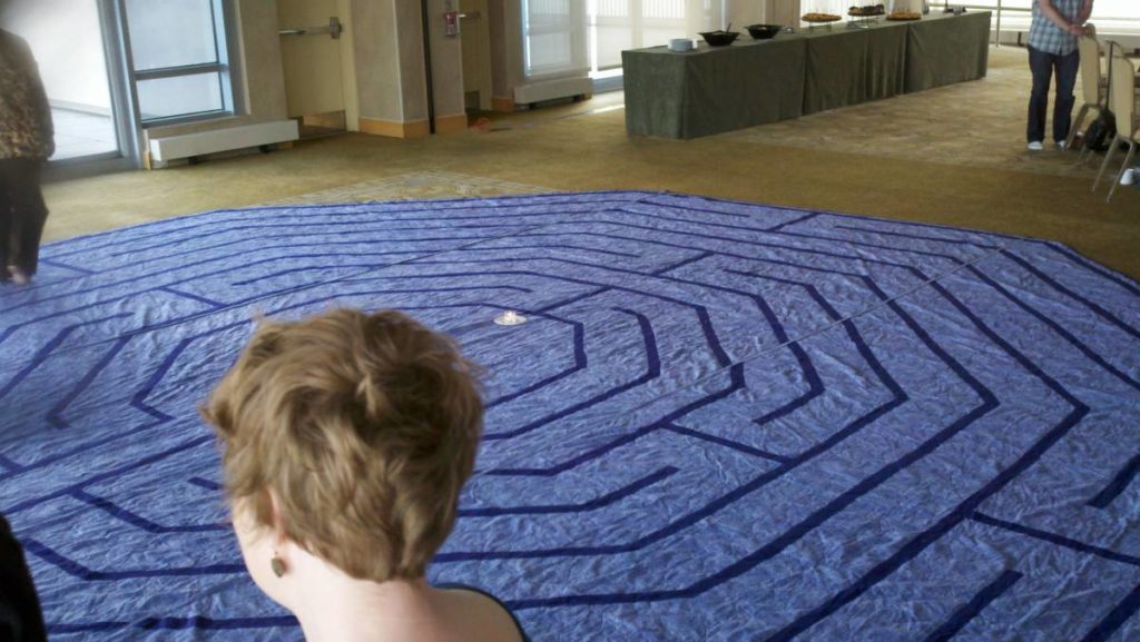 Photo shows a blue rug with a labyrinth design spread out on the floor for people to walk on.