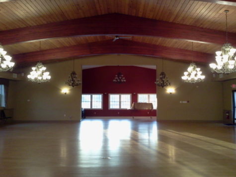 An empty ballroom. Chandeliers hang in two rows along the room. A Window lights the end of the room.