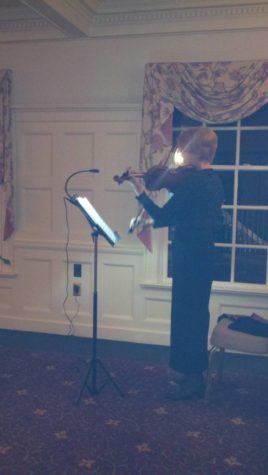 A violinist plays. Before her, a music stand is lit by a small light plugged into a nearby outlet. She's standing, but there's a chair waiting for her.