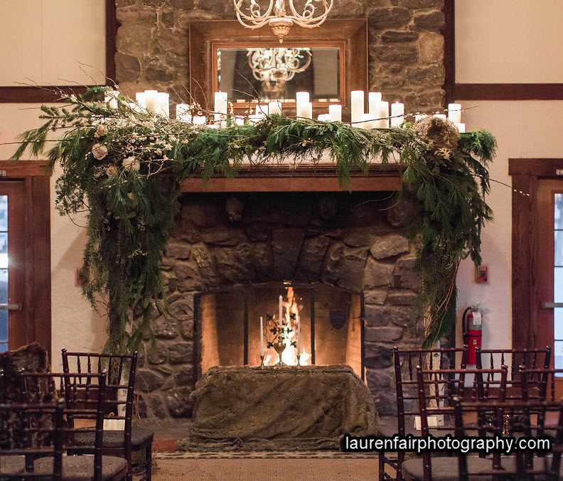 A huge stone fireplace with a roaring fire is framed by a wedding canopy of pine boughs. Beneath the canopy is a table with 3 candles and a chalice.