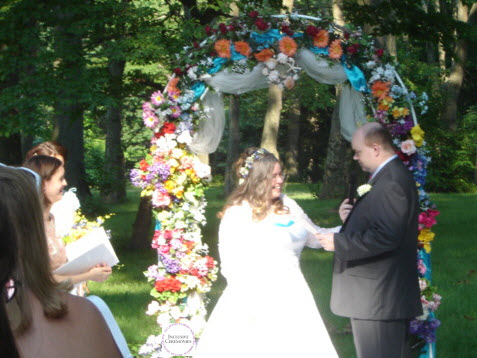 A summer wedding. The groom makes his vows. Bride and groom stand beneath a flowered arch.