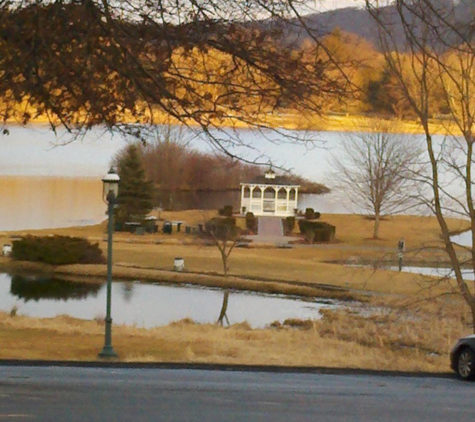 A gazebo at a golf course sits in the distance. A long grassy walk leads to the entrance and there is a lake behind it and water to either side of the walkway.