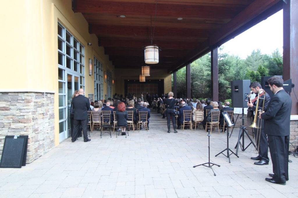 An intimate wedding space in a covered patio. Seats on either side line the aisle and a raised stage creates the ceremony space. Musicians play behind the guests with large speakers on stands facing the ceremony space.