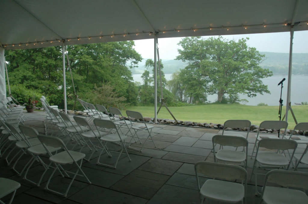 A large tented space overlooks a lake. White folding chairs are set up for a wedding ceremony and a microphone marks where the couple will stand.