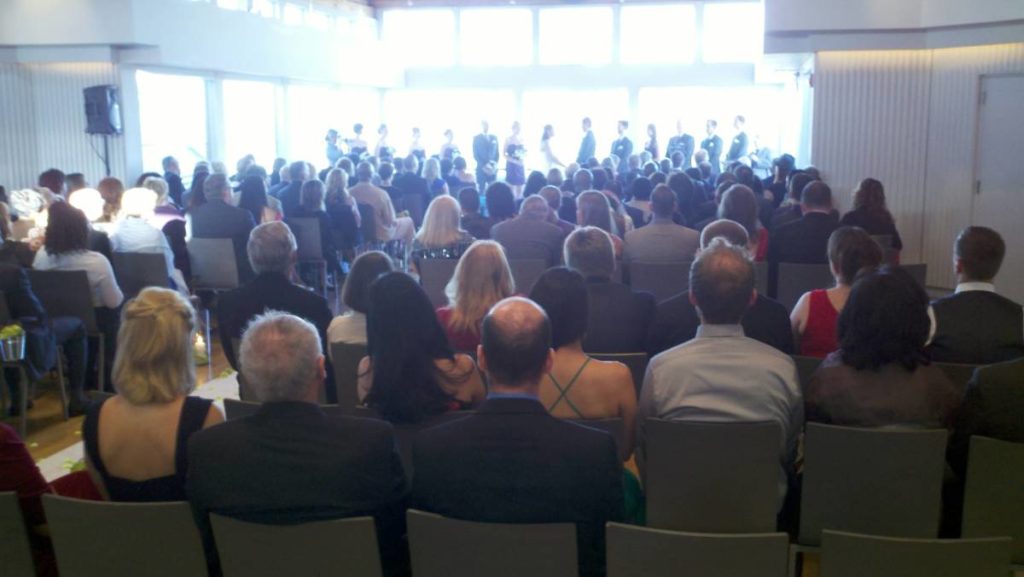 View of a wedding ceremony from behind the seated guests. The room narrows toward the front with wedding party in an alcove. Some guest seats face a small wall on either side of the alcove. 